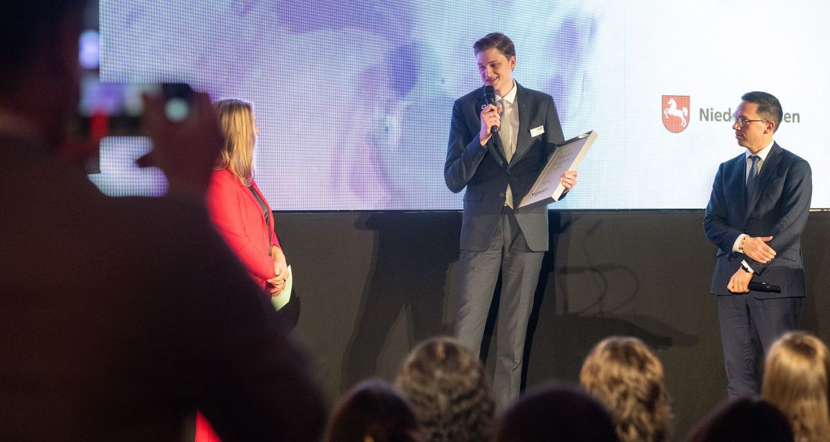 Jannes Dirks, Student der Jade Hochschule, wurde jetzt mit dem Niedersächsischen Wissenschaftspreis ausgezeichnet. Zu sehen ist er auf einer Bühne in Hannover, er hält die Preisurkunde und ein Mikrofon, neben ihm steht der niedersächsische Wissenschaftsminister Falko Mohrs (SPD).