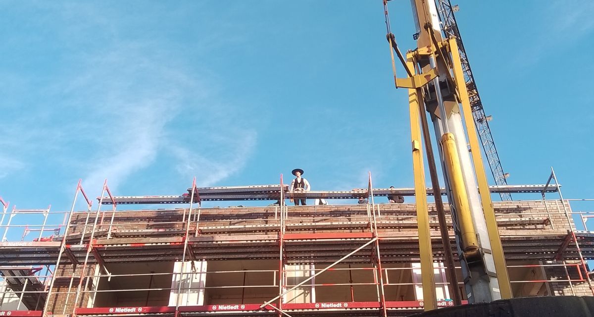 Ein Zimmermann steht beim Richtfest des Erweiterungsbaus der Oberschule Stadtmitte auf dem eingerüsteten Gebäude und spricht die Grußformel. Rechts im Bild ist ein gelber Kran zu sehen.