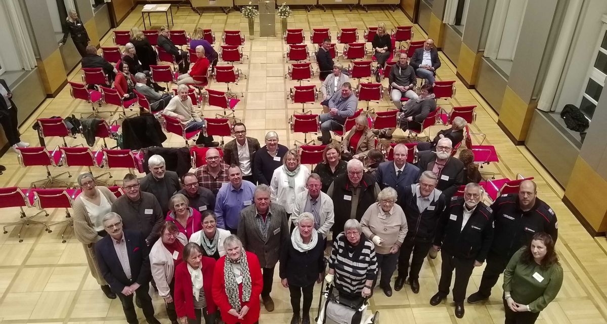 Im Ratssaal in Wilhelmshaven sind die Ehrenamtlichen zu sehen, die im Jahr 2024 für ihr Engagement geehrt werden. Eine Gruppe Personen steht vor aufgereihten roten Stühlen und schaut zur Zuschauerempore hinauf.