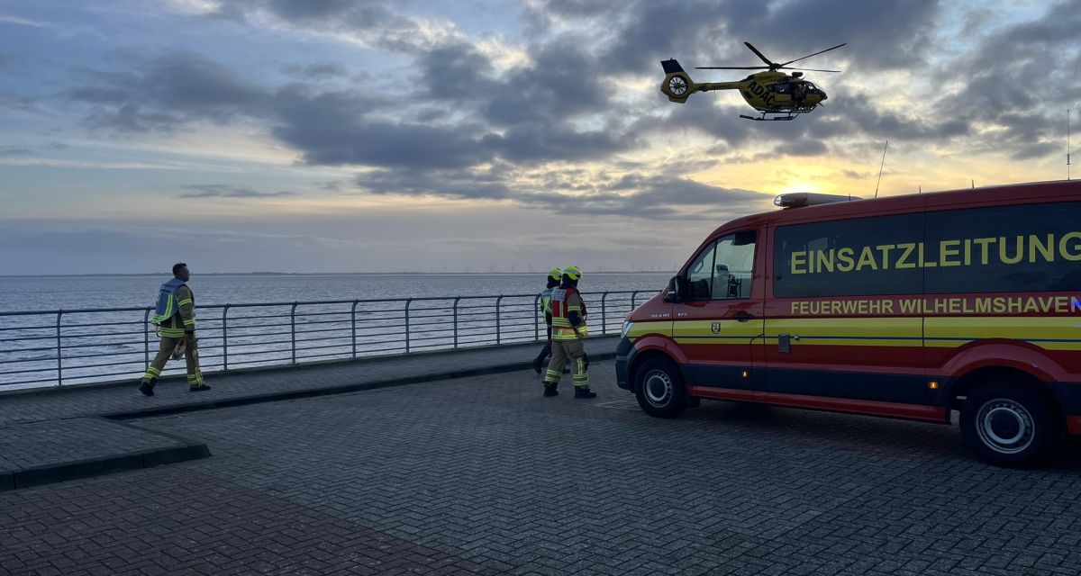 Zu sehen sind drei Feuerwehrleute in der Abenddämmerung am Fliegerdeich in Wilhelmshaven. Auf der rechten Seite des Bildes steht ein Einsatzleitungsfahrzeug der Feuerwehr Wilhelmshaven, über der Szenerie schwebt ein Luftrettungs-Hubschrauber des ADAC.