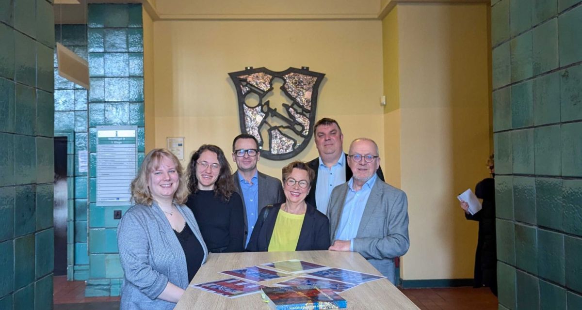 Zu sehen sind Mitglieder der Stadtverwaltung und die Nachfahren vom Künstler Georg Schmidt Westerstede vor einem Glasmosaikwappen der Stadt Wilhelmshaven von Westerstede.