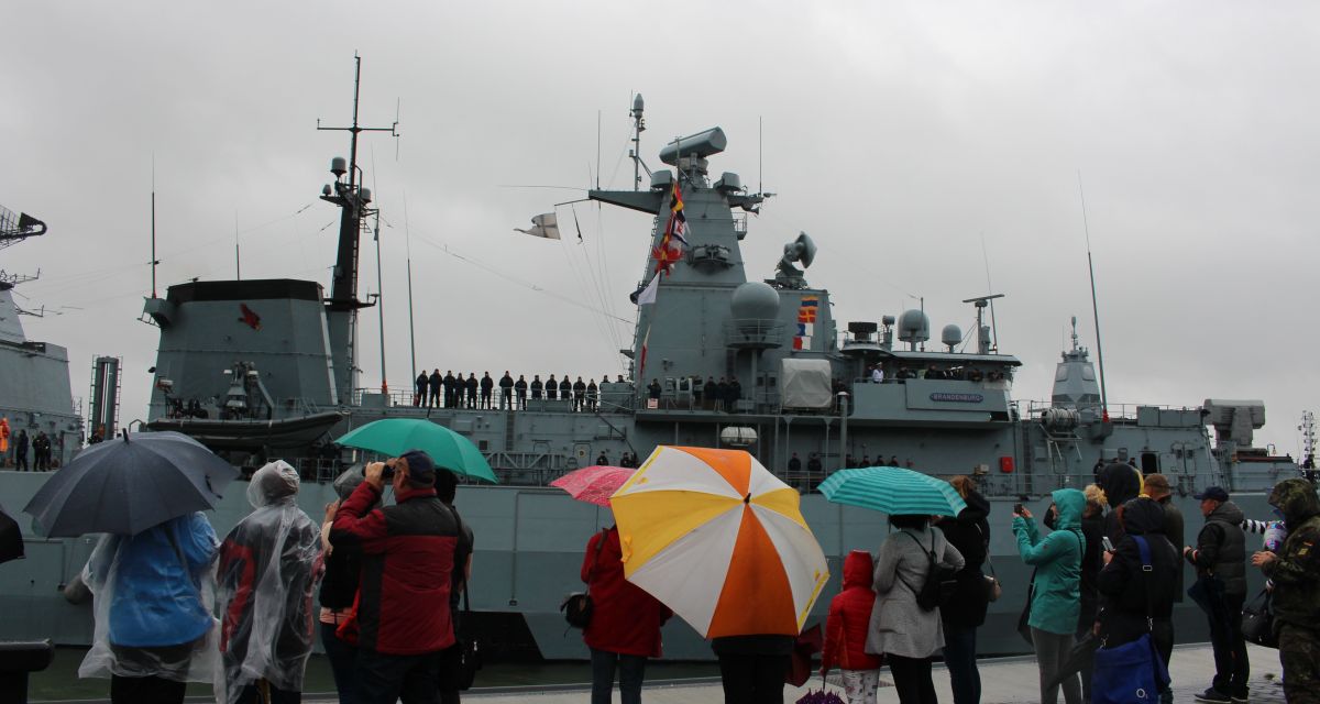 Zu sehen ist die Fregatte Brandenburg, davor Angehörige mit Regenschirmen. 