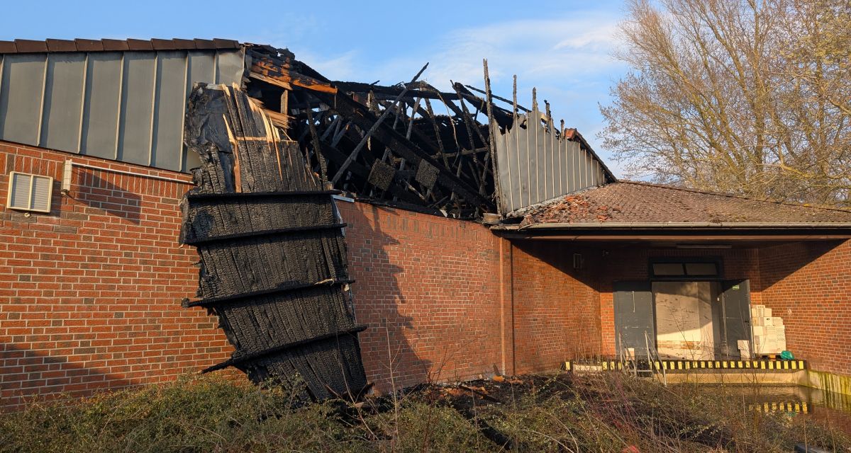 Zu sehen ist eine ausgebrannte Halle an der Güterstraße in Wilhelmshaven. Im Dachstuhl des Gebäudes klafft ein Loch, aus dem verkohlte Holzbohlen herausragen. Der Giebel des Dachs hängt verbogen und verkohlt herunter.