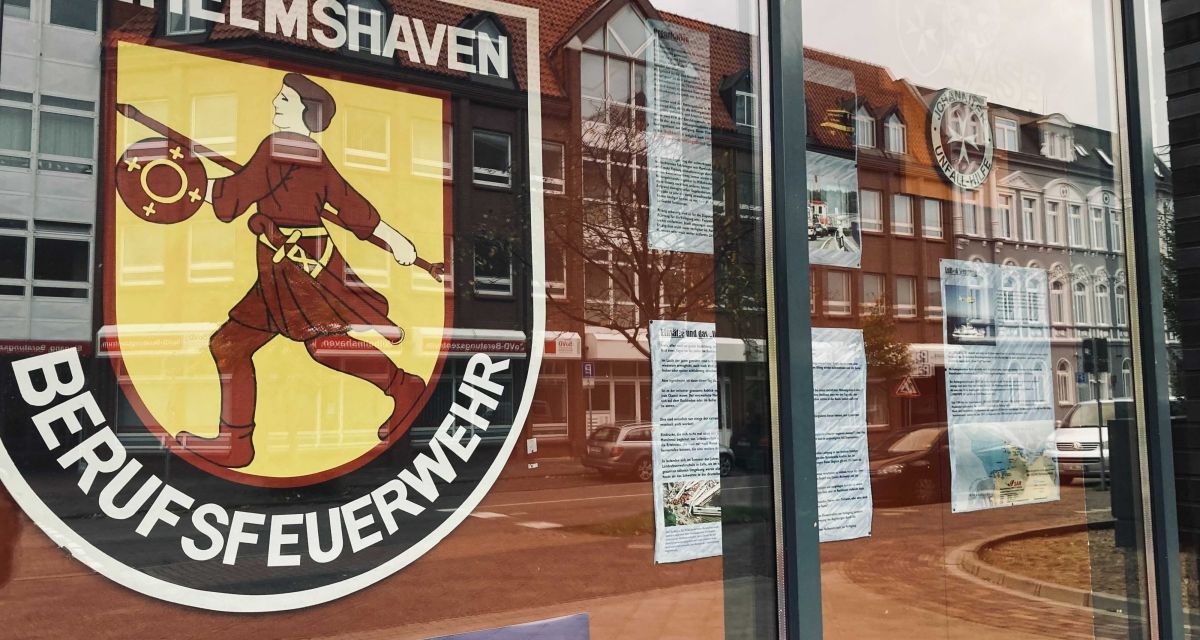 Ein Schaufenster an der Hauptwache der Berufsfeuerwehr Wilhelmshaven. Zu sehen ist das Wappen der Feuerwehr und Erklärungen zur Arbeitsweise und Ausrüstung.