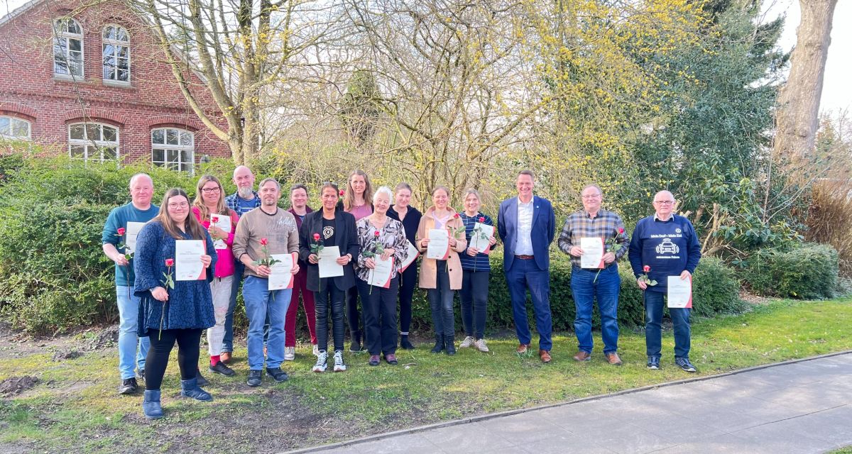 Zu sehen sind die frisch ausgezeichneten Dorfmoderatorinnen und -moderatoren für den Landkreis Wittmund und die Inseln gemeinsam mit Wittmunds Landrat Holger Heymann. Sie halten jeweils eine Urkunde und eine rote Rose in Händen.
