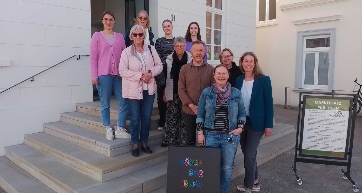 Die Reihe für Demokratie im Jahr 2025 wird in der Börse der Ideen in Varel vorgestellt. Auf der Haupttreppe der Börse stehen (von links nach rechts): Fenja van Düllen (Gertrud und Hellmut Barthel Stiftung), Sigrid und Karin von Omas gegen Rechts Varel, Liv Blendermann (FSJlerin der Barthel-Stiftung), Iris von den Omas gegen Rechts Varel, Leena Küchel (FSJlerin der Agenda Varel), Thomas Dewald von der Ambulanten Wohnungsnotfallhilfe der Diakonie, Anne Büttner (Geschäftsführerin Agenda und Freiwilligenagentur Varel, Wiebke Bartels (Ambulante Wohnungsnotfallhilfe und Janita Budde-Frerichs (Vernetzerin Agenda Varel). Vor ihnen steht ein Schild mit der bunten Aufschrift