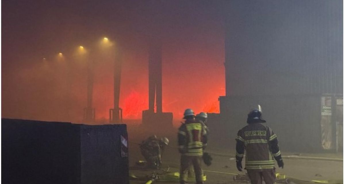 Ein Brand im Abfallwirtschaftsbetrieb Wiefels im April 2025. Zu sehen sind vier Feuerwehrleute in Einsatzkleidung und mit Helmen, die eine Löschleitung legen. Im Hintergrund lodert orange-rot das Feuer in einer Lagerhalle, Rauch füllt die Luft.
