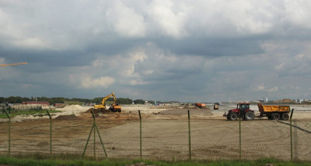 Die Bauarbeiten auf dem Flugplatz Wittmundhafen. Hinter einem Zaun sind Baumaschinen auf sandiger Fläche zu sehen, im Hintergrund ist die Start- und Landebahn ebenso zu erkennen wie einzelne Gebäude.