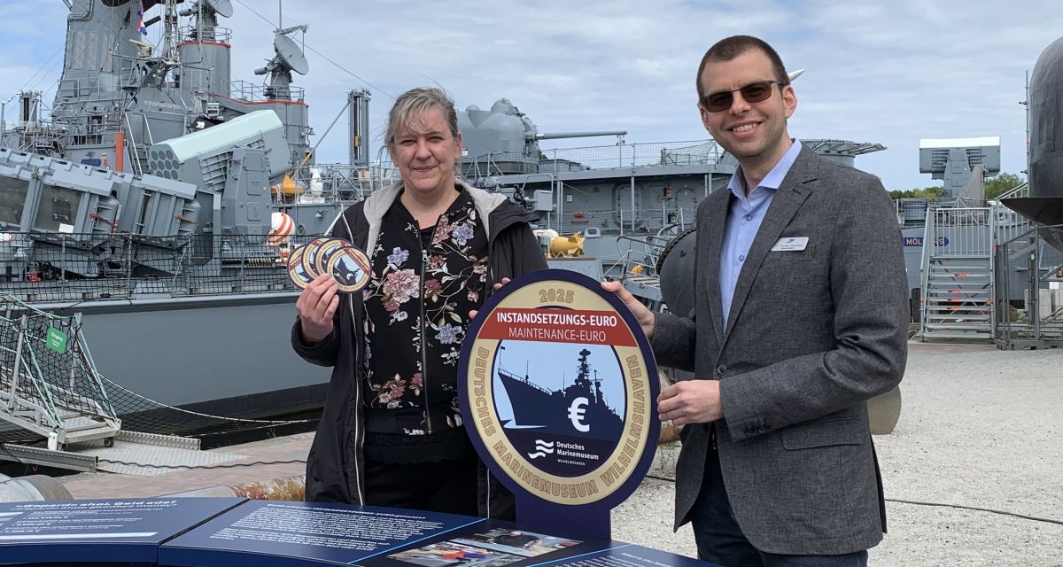 Auf dem Foto sind der Leiter des Marinemuseums, Dr. Thomas Eisentraut (rechts), und seine Stellvertreterin, Nina Nustede (links), zu sehen. Auf dem Außengelände des Marinemuseums werden Informationstische aufgebaut. Dort wird über die Kosten für die Instandhaltung oder -setzung der Schiffe informiert. Mit dem Instandsetzungs-Euro werden so Spenden für diese Aufgaben gesammelt. Im Hintergrund des Bildes sind einige der Museumsschiffe zu sehen. 