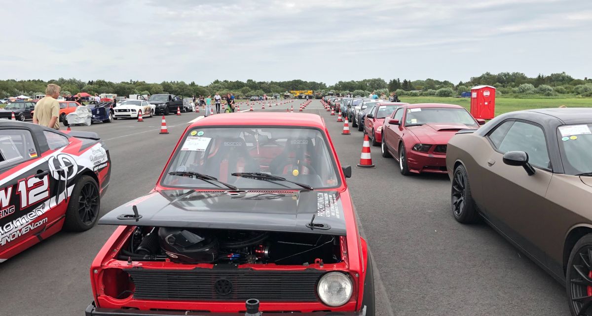 Mehrere verbastelte Autos auf einem Flugfeld vor dem Start eines Drag races