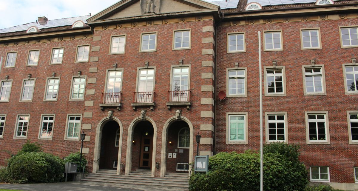 Zu sehen ist das Kreisamt des Landkreises Friesland in Jever. Der Bau ist in rotem Klinker mit grau abgesetzten Fenstern und Ecksteinen gehalten. Leicht nach vorn gesetzt ist der Eingangsbereich, über dem ein Adler im Giebel prangt. Über drei kleineren Torbögen ist jeweils ein steinerner Kopf angebracht.