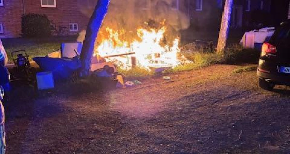 Zu sehen ist ein brennender Haufen Sperrmüll auf einer Freifläche an der Paul-Hug-Straße in Wilhelmshaven. Das Feuer war in den frühen Morgenstunden von Dienstag, 29. Juli, ausgebrochen. Im Hintergrund sind rotgeklinkerte Wohnhäuser zu erkennen, am linken Bildrand steht ein Feuerwehrmann, am rechten Bildrand ein schwarzer Pkw. Die Bäume am Rand des Bürgersteigs sind durch die Signallichter der Feuerwehr blau erleuchtet.