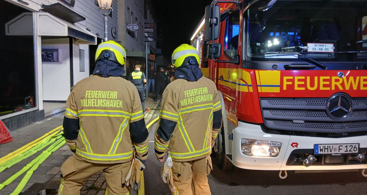Zwei Feuerwehrleute der Feuerwehr Wilhelmshaven stehen in Einsatzkleidung vor dem Gebäude einer ehemaligen Diskothek in der westlichen Marktstraße in Wilhelmshaven. Die Einsatzkräfte haben der Kamera den Rücken zugedreht, rechts neben ihnen ist ein Löschfahrzeug mit ausgerollten Schläuchen zu sehen, vor ihnen unter anderem ein Beamter der Polizeiinspektion Wilhelmshaven/Friesland. Anlass des Einsatzes war ein Feuer im Gebäude, das in der Nacht Ende Juli schnell gelöscht werden konnte.