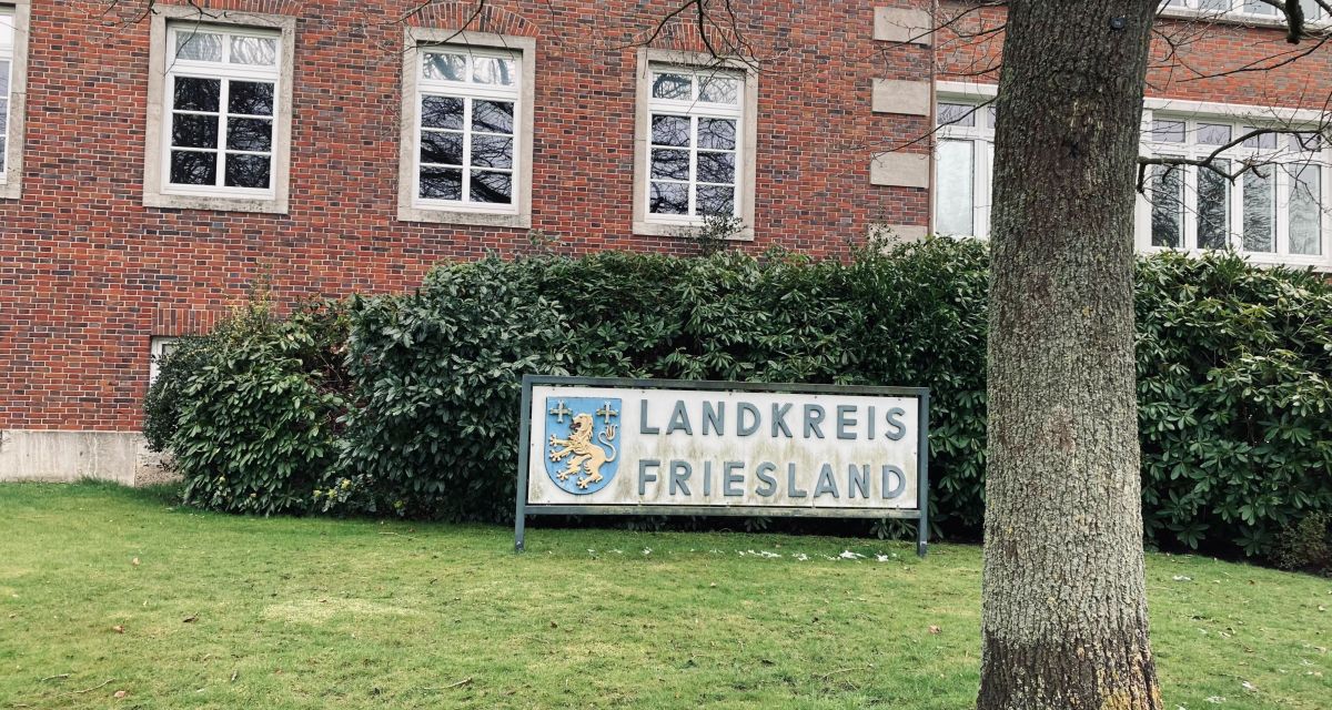 Ein Schild vor dem Kreisamt des Landkreises Friesland in Jever: links auf dem weißen Schild ist das Wappen des Landkreises zu sehen, ein gelber Löwe zwischen zwei goldenen Kreuzen auf blauem Grund. Neben dem Wappen steht in blauen Buchstaben 