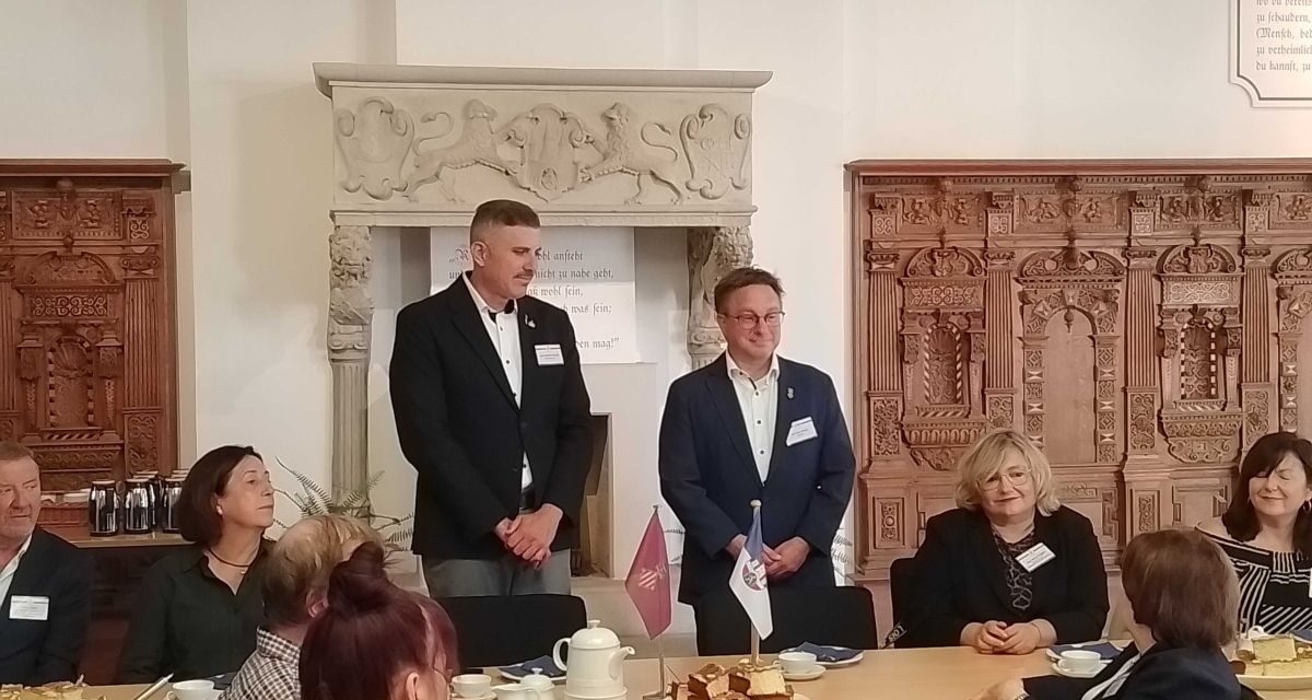 Der Bürgermeister der Stadt Jever, Jan Edo Albers, begrüßt die Delegation aus Jevers spanischer Partnerstadt Cullera im Graf-Anton-Günther-Saal des jeverschen Rathauses. Neben Albers steht sein Amtskollege aus Cullera, Jordi Mayor Vallet. Vor ihnen sind die Stadtflaggen der beiden Städte aufgestellt, im Hintergrund ist ein Kamin und eine verzierte Holzverkleidung zu sehen. Um die beiden Bürgermeister herum sitzen Vertreter*innen der Verwaltungen von Jevr und Cullera, auf den Tischen stehen Teekannen und Teekuchen.
