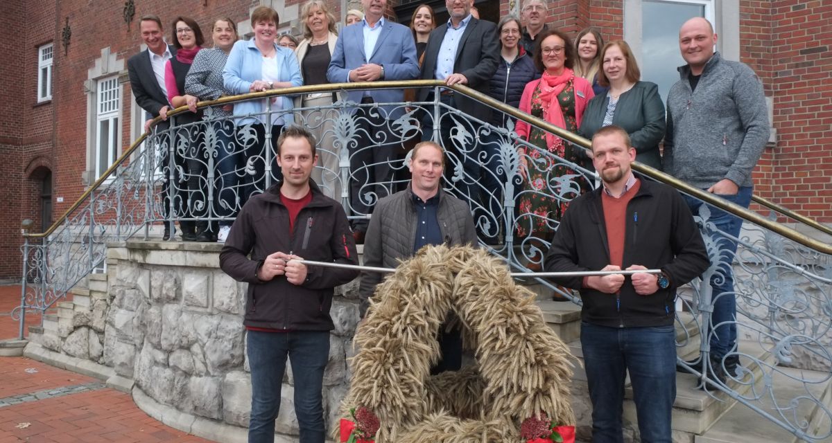 Übergabe der Erntekrone im Kreis Wittmund. Auf und vor der Treppe des Kreishauses stehen Vertreter*innen von Landwirtschaft, Politik und der KreisLandFrauen. Im Vordergrund ist die traditionelle Erntekrone aus Ähren mit roten Schleifen zu sehen.