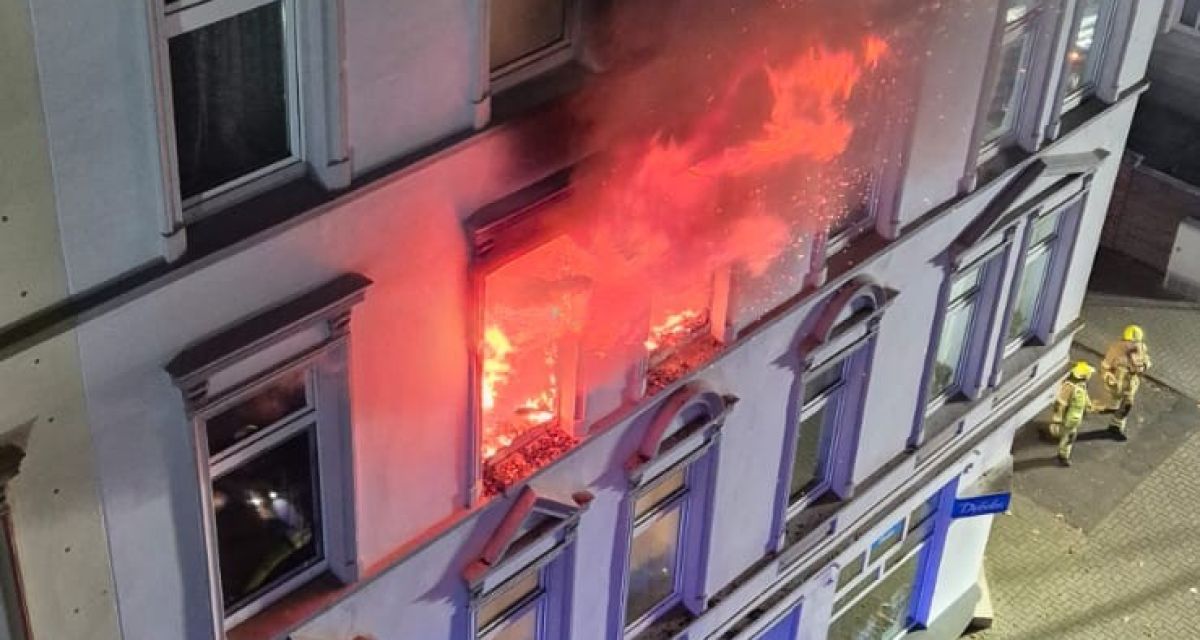 Zu sehen ist ein Brand in einer Wohnung in der Mitscherlichstraße in Wilhelmshaven. Aus dem Korb eines Einsatzfahrzeuges der Feuerwehr zeigt das Bild das Gebäude, aus Fenstern im zweiten Obergeschoss schlagen Flammen und Rauch. Am Boden sind ein Löschwasserschlauch, Schutt und eine Feuerwehraxt zu sehen. An der Ecke des Gebäudes stehen zwei Feuerwehrleute in gelber Schutzkleidung.