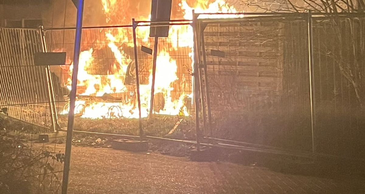 Zu sehen ist der Brand zweier Fahrzeuge in der Nacht zum 28. Dezember 2025 in der Marktstraße in Wilhelmshaven. Hinter zahlreichen Baustellenzäunen lodern in der Dunkelheit Flammen und Rauch aus nur undeutlich zu erkennenden Autos.