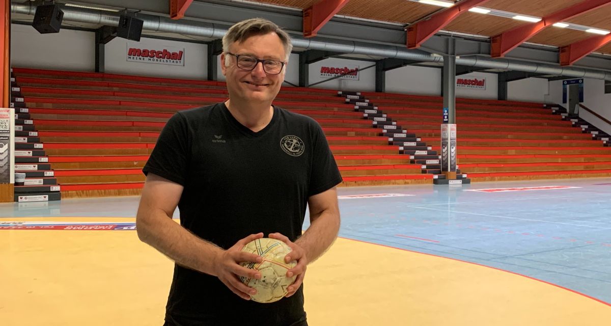 Zu sehen ist Arek Błacha. Er ist der Trainer der Handballspielgemeinschaft Varel (HSG Varel). Er steht in der Manfred-Schmidt-Sporthalle in Altjührden, die heimische Halle des Vereins. Er hält einen Handball in der Hand und trägt ein schwarzes T-SHirt mit dem Vereinslogo. 