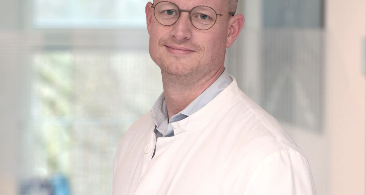 Zu sehen ist der neue Chefarzt der Neurologie an den Friesland Kliniken, Dr. Lambert Schless. Er trägt kurzgeschnittene Haare, eine runde Brille und einen weißen Arztkittel.