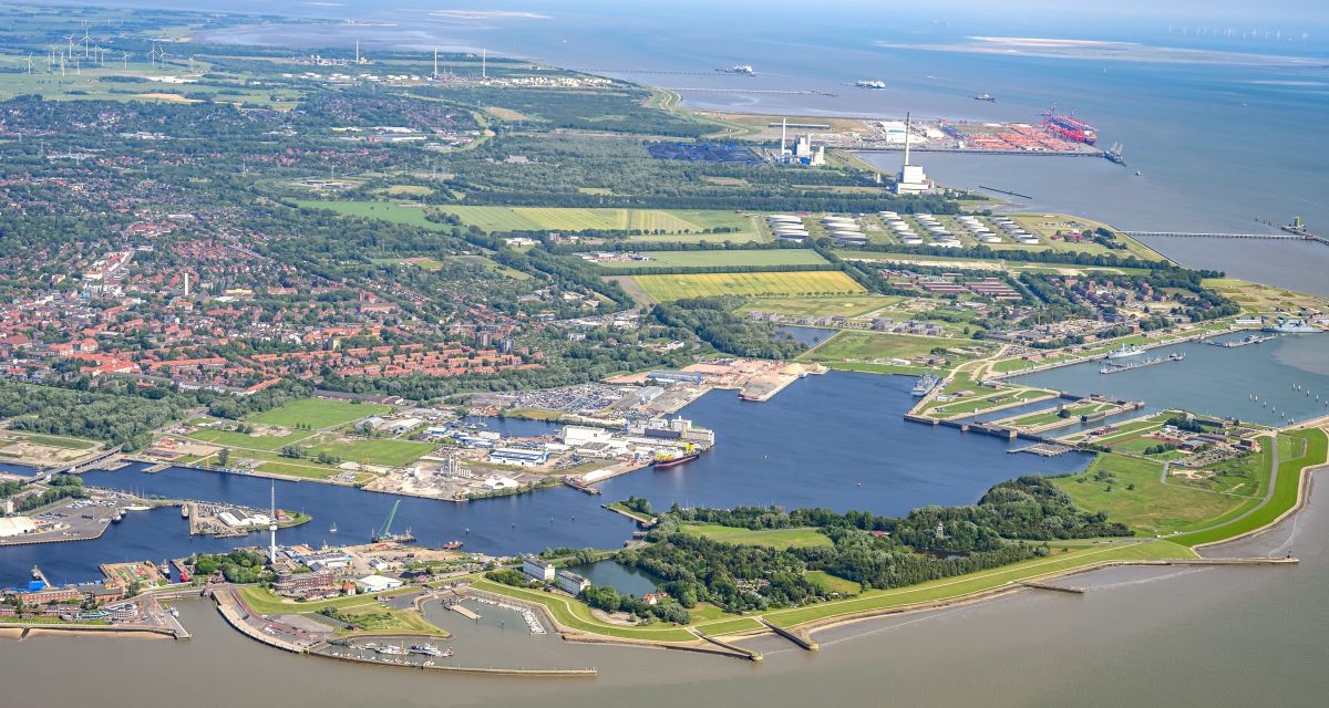 Zu sehen ist der Innere Hafen in Wilhelmshaven. Am linken Bildrand ist die Jachmannbrücke und darüber die Wohnbebauung der Stadt zu erkennen, rechts die Seeschleuse und im Hintergrund der Marinestützpunkt Heppenser Groden und der JadeWeserPort.