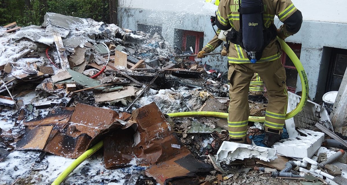 Zu sehen sind zwei Feuerwehrleute der Feuerwehr Wilhelmshaven, die einen Brand auf dem Hof eines Mehrfamilienhauses in der Ebertstraße in der Südstadt gelöscht haben. Sie stehen inmitten eines Haufens an Umrat und Bauschutt, der teilweise mit Löschschaum durchsetzt ist. Ein Löschschlauch schlängelt sich durch die Brandmasse.
