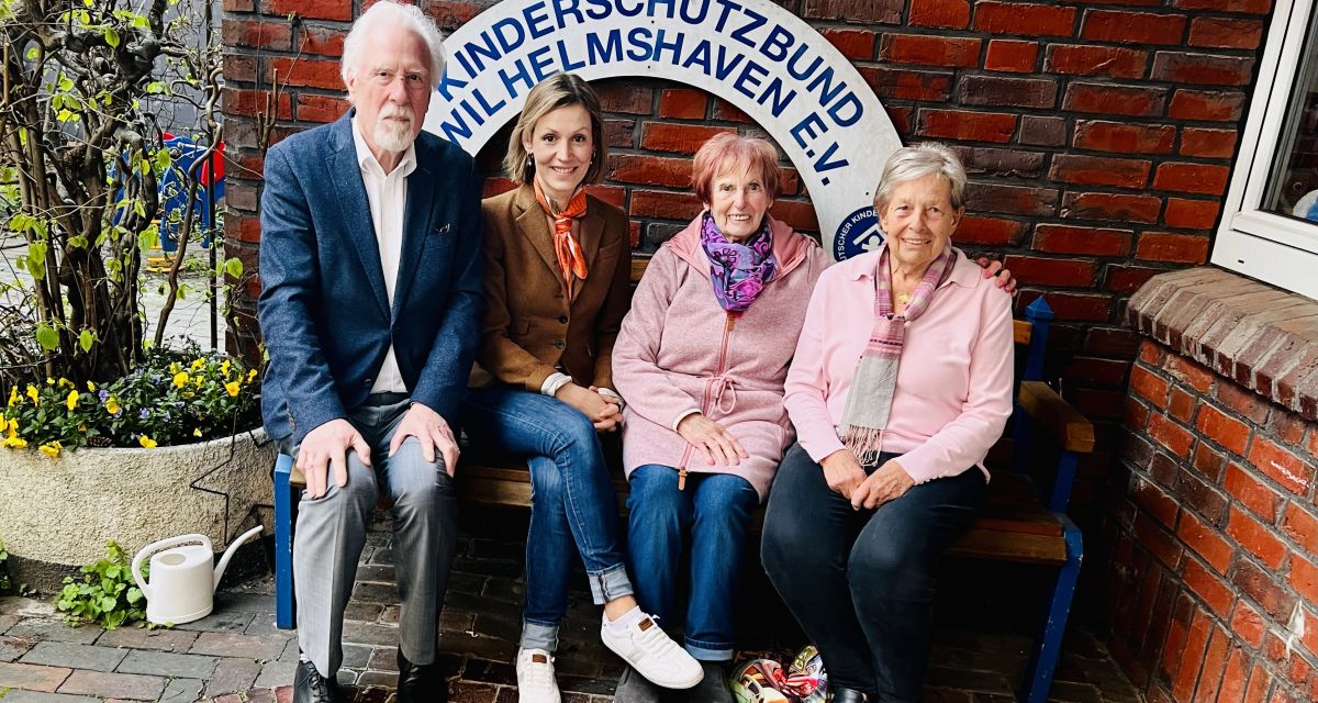 Zu sehen ist der im Jahr 2026 neu gewählte Vorstand des Ortsvereins Wilhelmshaven des Deutschen Kinderschutzbundes. Von links nach rechts sitzen auf einer Bank vor einer roten Klinkerwand und einem weißen Rundbogen mit der Aufschrift 