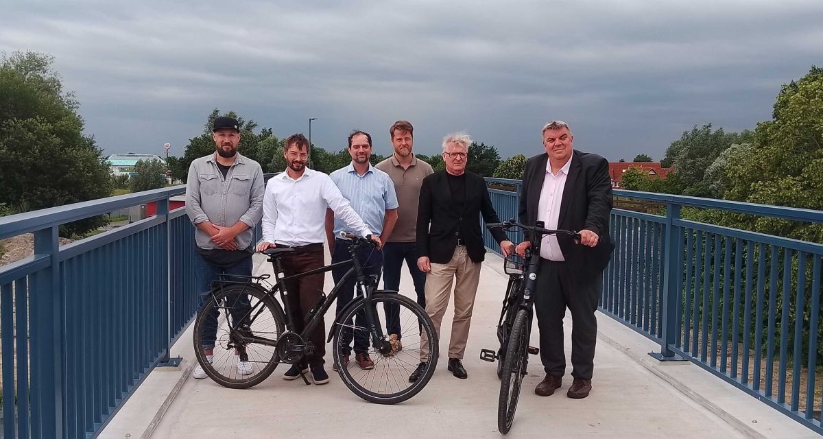Die Fahrrad- und Fußwegbrücke in Potenburg wird offiziell eröffnet. Auf der Brücke über die Hooksieler Landstraße, deren Geländer im bekannten Eisenglimmerblau gestrichen ist, stehen (von links nach rechts) Marius Wilke, André Lachmund, Martin Heinß, Karsten Baus, Tim Menke und Oberbürgermeister Carsten Feist. Lachmund und Feist haben Fahrräder dabei.