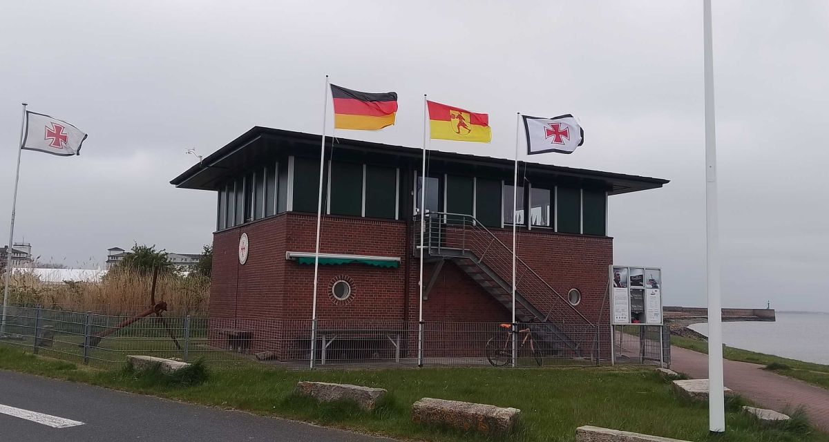 Zu sehen ist das Stationsgebäude der Deutschen Gesellschaft zur Rettung Schiffbrüchiger am Helgolandkai in Wilhelmshaven. Das mehrstöckige Klinkergebäude hat ein Obergeschoss mit großen Fenstern. Vor dem Gebäude wehen die schwarz-rot-goldene Deutschlandflagge, die rot-gelbe Wilhelmshavener Stadtflagge mit dem Stadtwappen des Rüstringer Friesen und die Flagge der Seenotretter mit schwarz umrandetem weißen Feld und darin einem roten Hansekreuz. Der Himmel ist bedeckt.