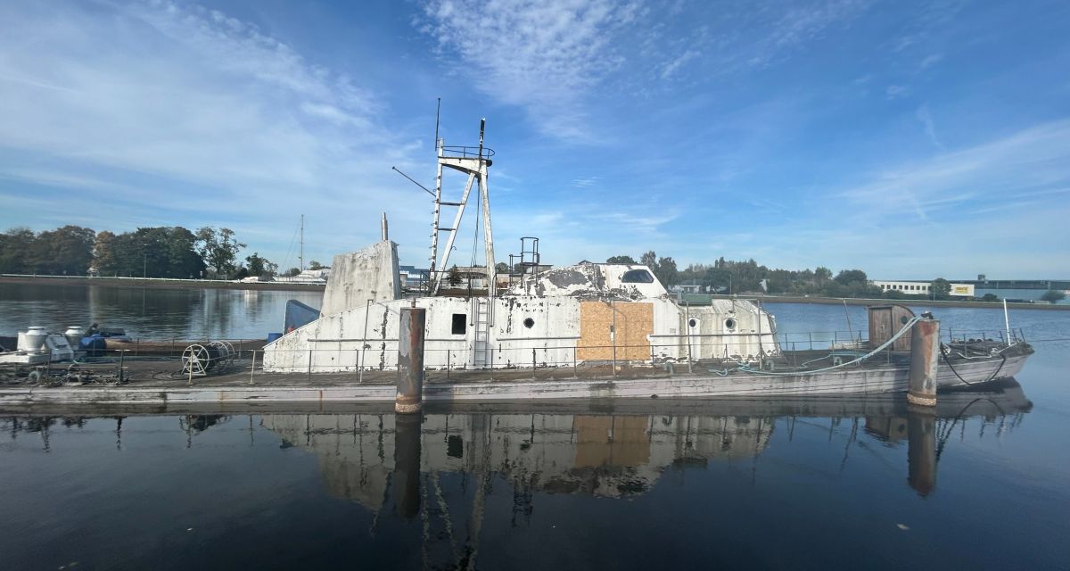 Man sieht ein Schiff von der Seite, wie es etwas schräg im Wasser liegt. Das Schiff sieht schon sehr beschädigt aus.