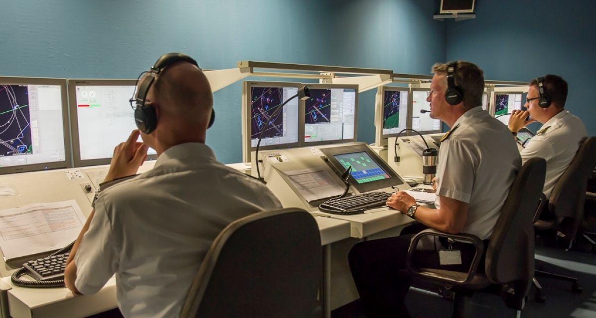 Drei Männer in kurzen weißen Hemden sitzen mit Kopfhörern vor einer Reihe von Bildschirmen.