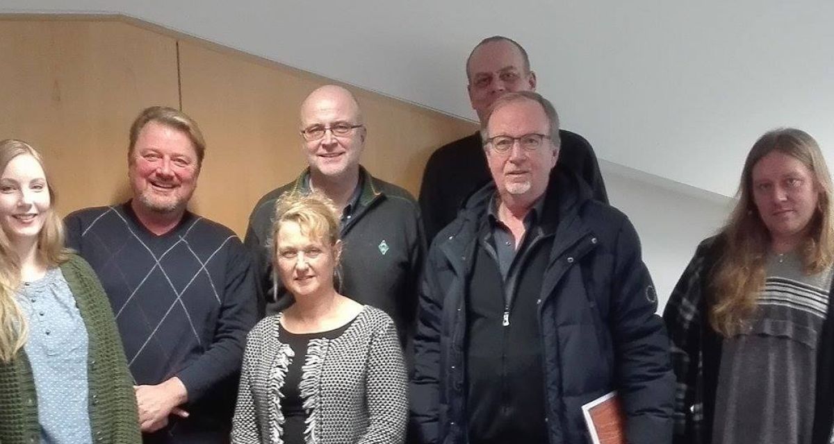 Sieben Vertreter aus Politik und Sozialem stehen vor einer holzvertäfelten Wand und schauen in die Kamera.