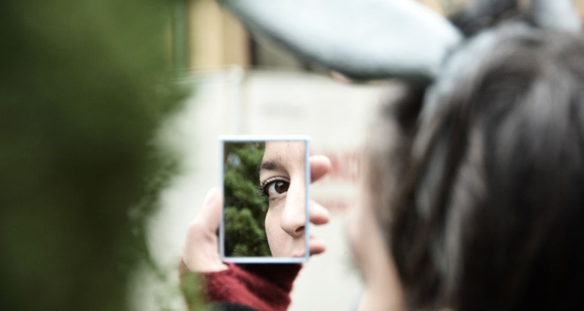 Eine junge Frau betrachtet sich im Spiegel. Zu sehen ist ein Teil ihres Hinterkopfs. Im Spiegel Nase und ein Auge.