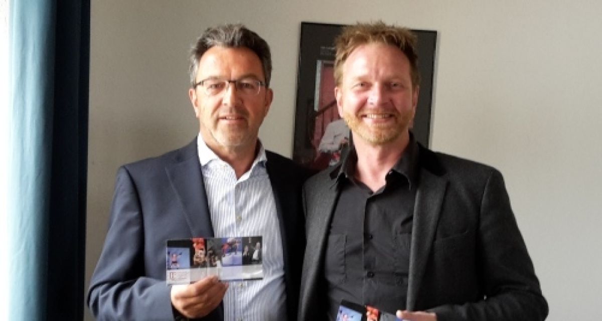 Zwei Männer lächeln in die Kamera. Sie haben Flyer in der Hand.