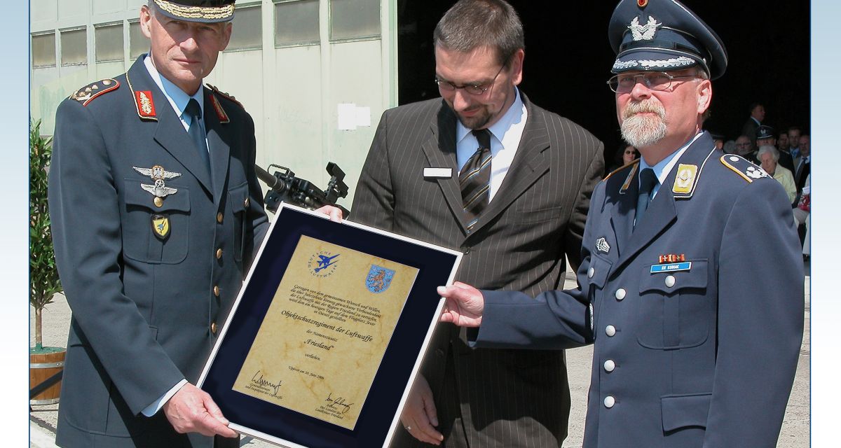 Zwei ranghohe Bundeswehrbedienstete und Frieslands Landrat Sven Ambrosy präsentieren für die Kamera eine eingerahmte Urkunde.
