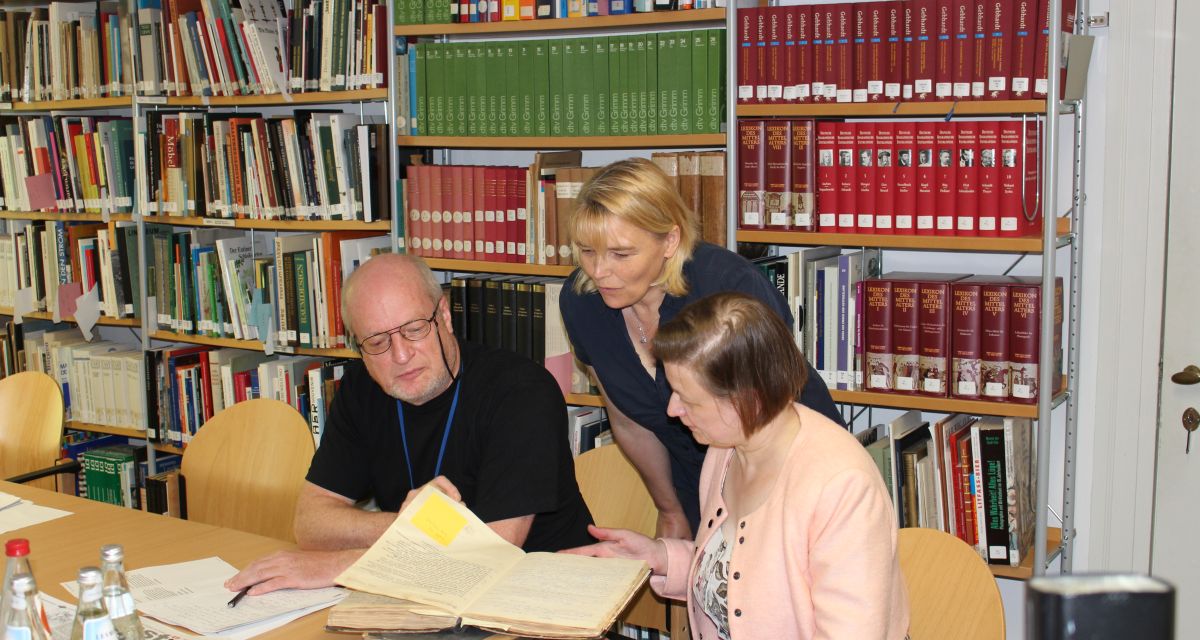 Vor einer mit Büchern gefüllten Regalwand sitzen zwei Personen, eine dritte steht, und blättern in einem Buch