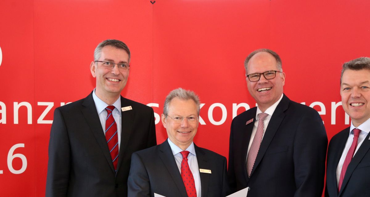 Vier Männer in schwarzen Anzügen und roten Krawatten vor einer roten Wand.