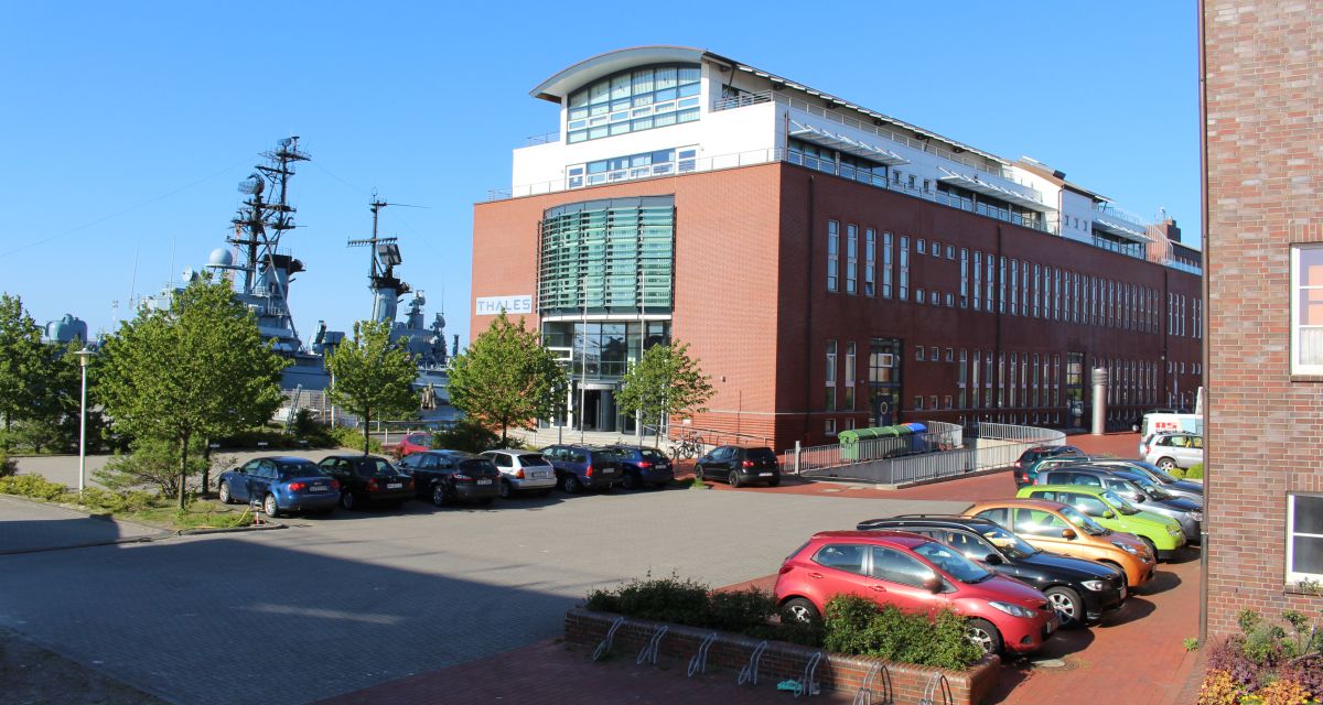 Zu sehen ist das Gebäude des Technologiekonzerns Thales mit Sitz in Wilhelmshaven gegenüber eines Marineschiffes.