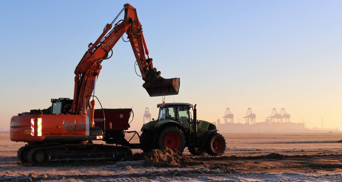 Zu sehen sind ein Traktor und ein Bagger auf einer weiten, leeren Fläche. 