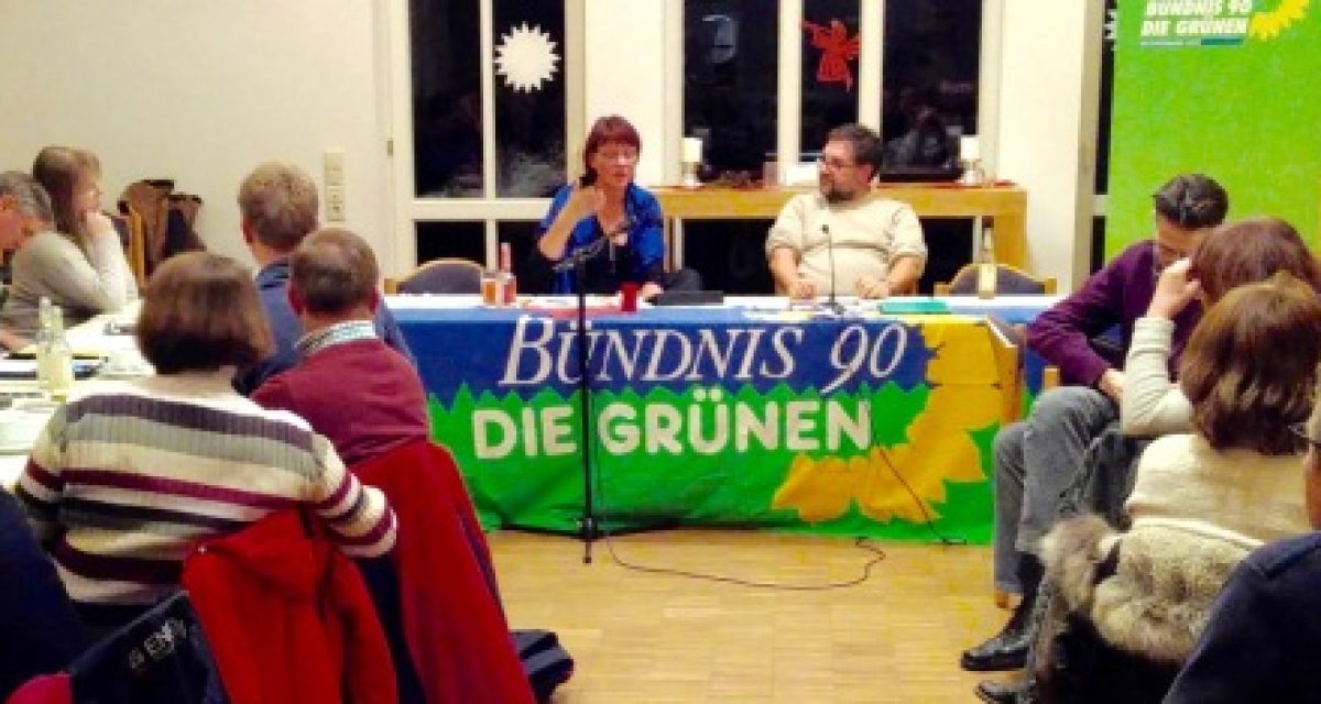 Ein Saal mit vielen sitzenden Menschen. In der Mitte ein Mann und eine Frau an einem Tisch auf dem ein blau-grünes Banner der Grünen liegt.