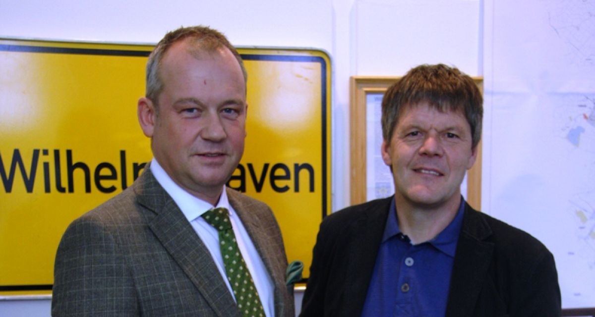 OB Wagner und Michael Diers lächeln in die Kamera. Im Hintergrund ein gelbes Ortsschild auf dem Wilhelmshaven steht. 