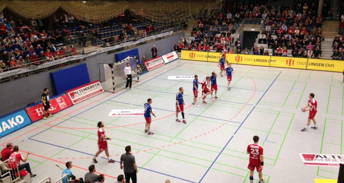 Handballspieler stehen auf dem Spielfeld. Publikum sitzt drumherum. 